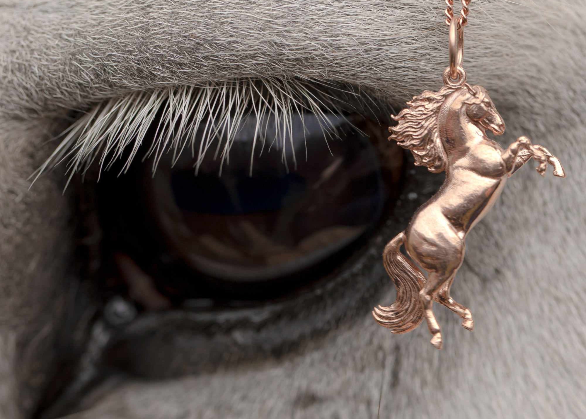 A beautifully crafted rose gold horse pendant, distinguished by its long tail and exquisite design. Our Rearing Wild Horse Pendant on a chain, highlighting the beauty of equestrian-inspired jewelry with details on its hair and tail. The necklace chain is also rose gold and has a series of small rounded beads interspersed. The Rose Gold Wild Horse Pendant is the best gifts for an equestrian, horse riding, horse girls, cowgirls and for horse enthusiasts.
A brown horse's neck in the background.
