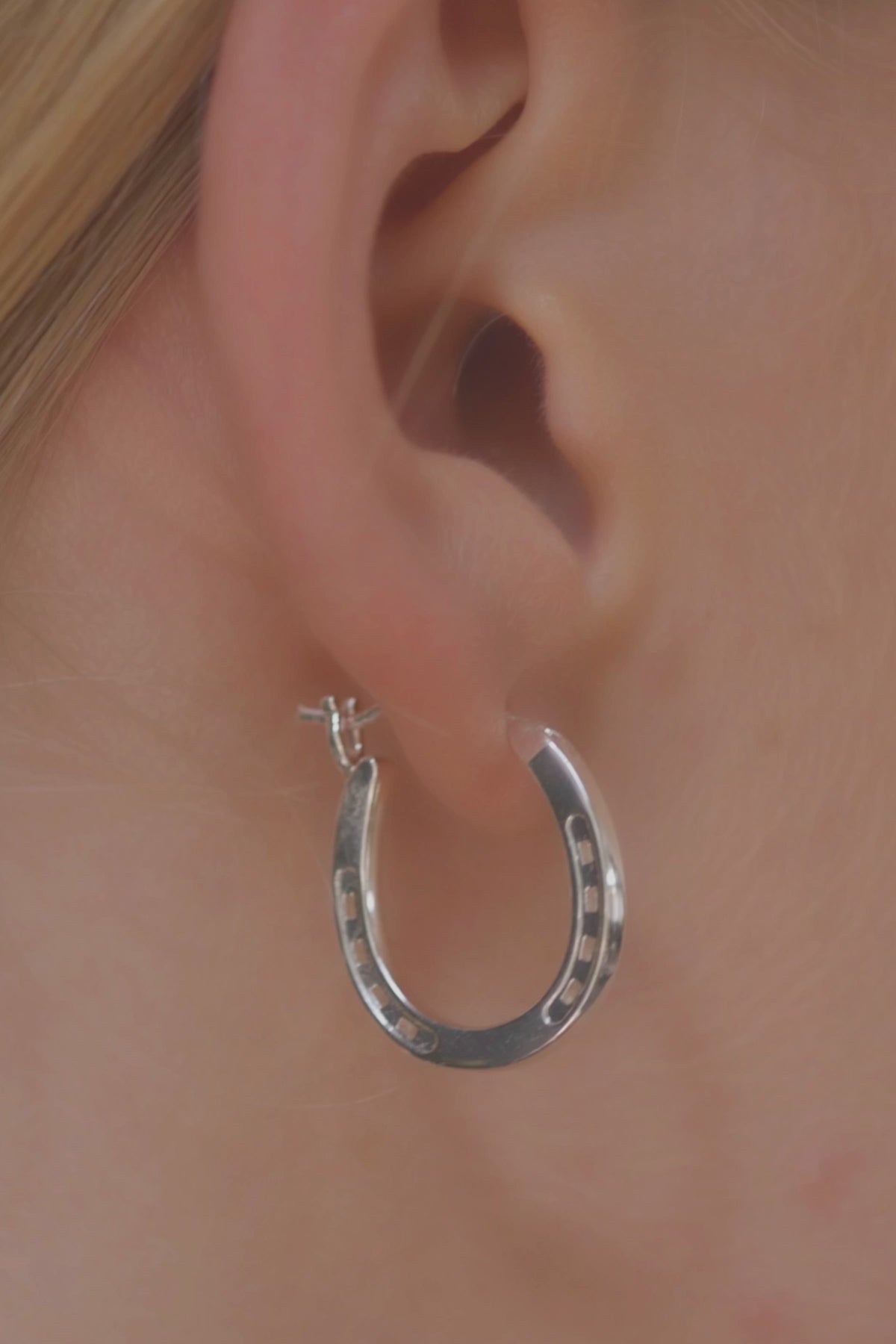 A close-up of a pair of elegant silver hoop horseshoe earrings on a blond model, emphasizing their gleaming finish and stylish form. Our silver Horse Shoe Earrings are unique equestrian gifts crafted from high-quality gold. This silver horseshoe earrings are the perfect equestrian lifestyle fan gift and a good luck charm. Our Horseshoe earrings are the perfect birthday present for horse lovers, horse girls, cowgirls. A representation of good luck.