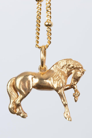 The Friesian Horse Pendant is a gold pendant in the shape of a horse, hanging from a gold chain. The FRIESIAN horse is depicted mid-stride with intricate details, including a flowing mane and tail. The chain features small, rounded beads spaced along its length. Ideal as a special equestrian gift for women, equestrians, horses and Friesian horses lovers.