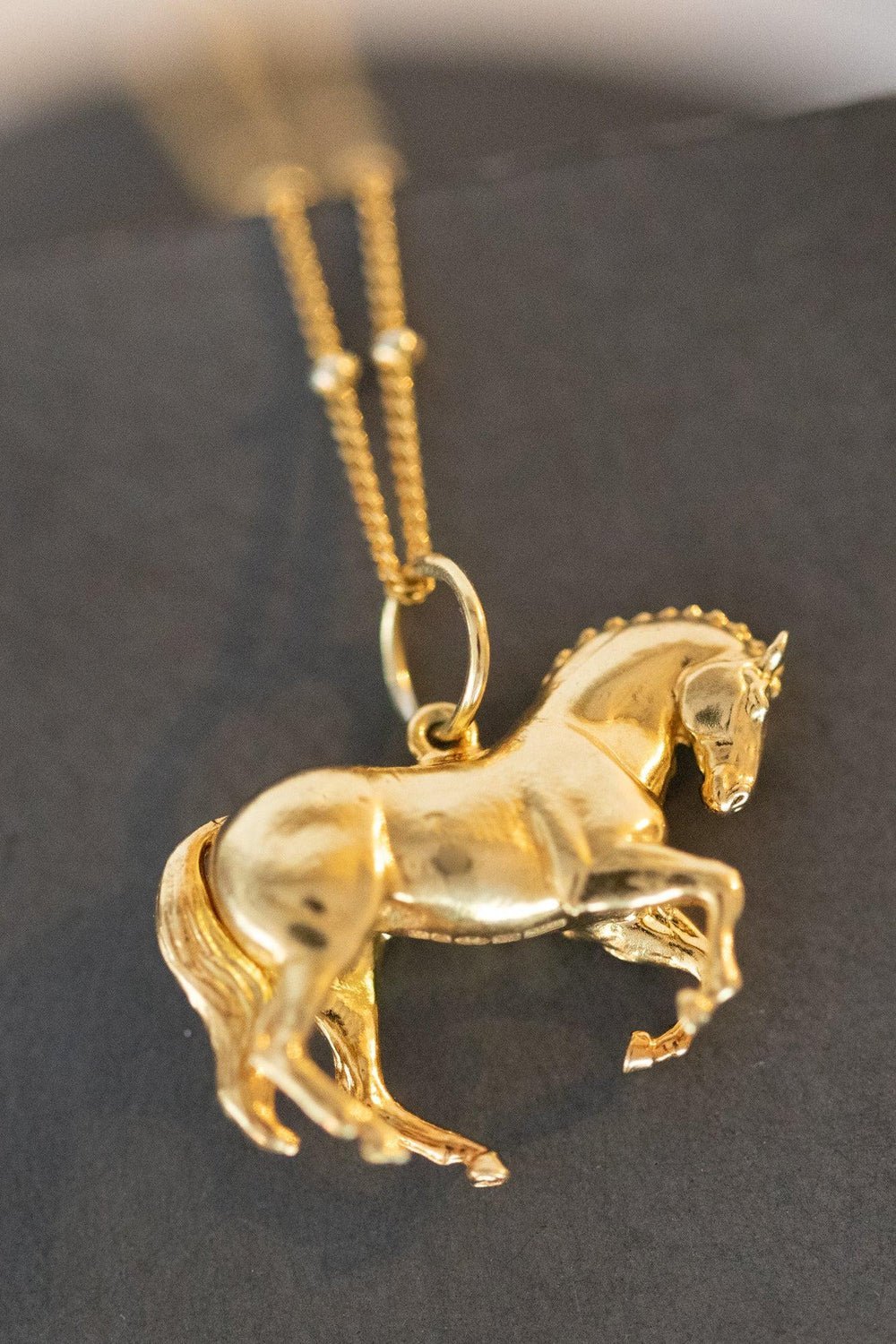 A beautifully crafted gold horse pendant hanging from a fine chain, highlighting its luxurious and artistic features.
The pendant is shaped like a  wild horse in mid-stride, with detailed features such as its mane and tail. The necklace chain is also gold and has a series of small, rounded beads interspersed along its length. The background appears to be a plain or stone , light-colored surface.  Perfect for horse lovers, fans of equestrian inspired jewelry, horseback riders,  gold horse jewelry.