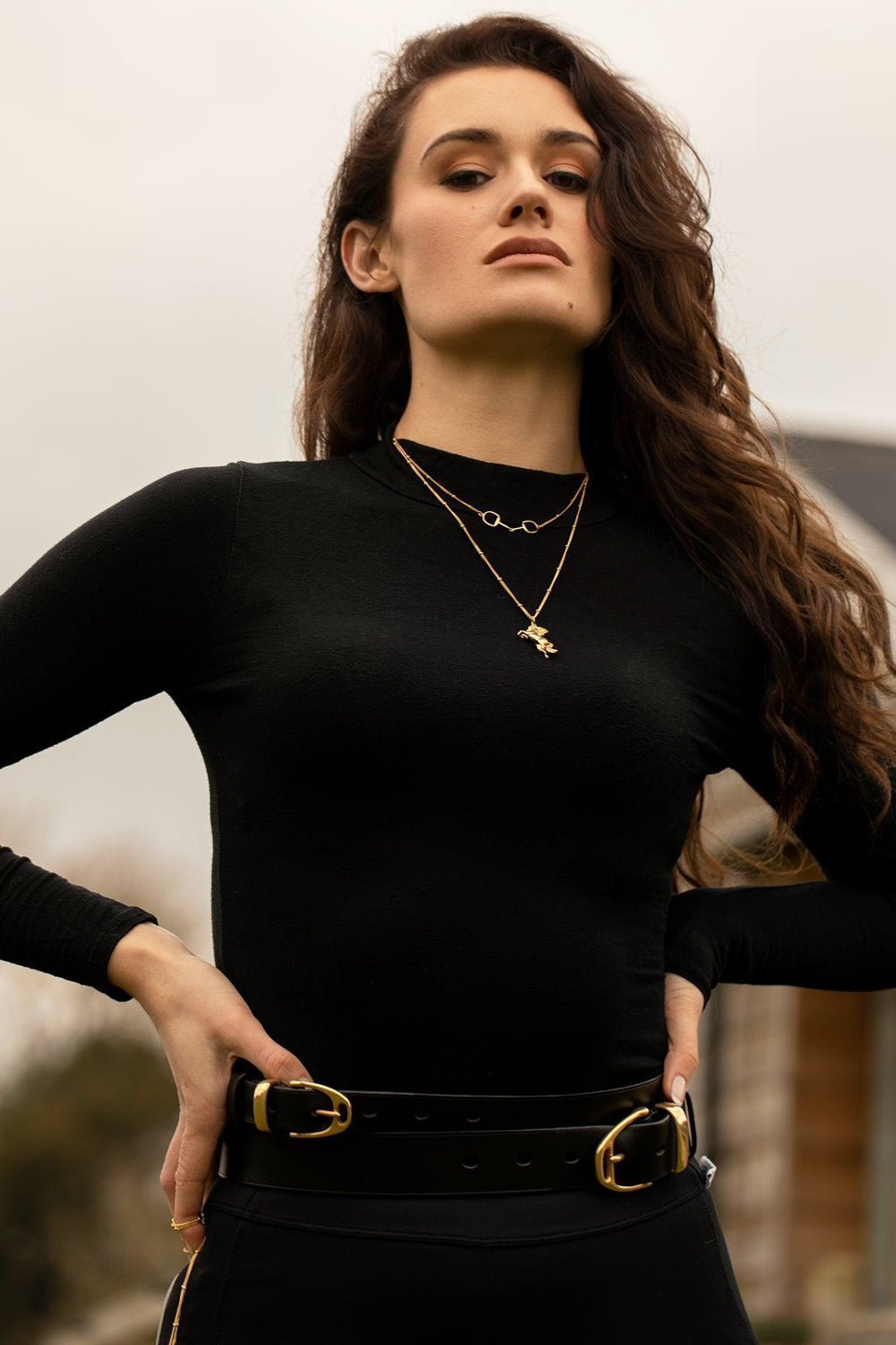 Brunet model  woman wearing equestrian outfit. She is wearing a stylish gold horse necklace with a layering of Snaffle Bit necklace. The necklace chain is also gold and has a series of small rounded beads interspersed. This Snaffle Bit pendant necklace is the perfect for horse enthusiasts and fashion lovers, it adds a refined touch to any outfit and makes a memorable equestrian gifts for women and horse themed gifts, gold girls.