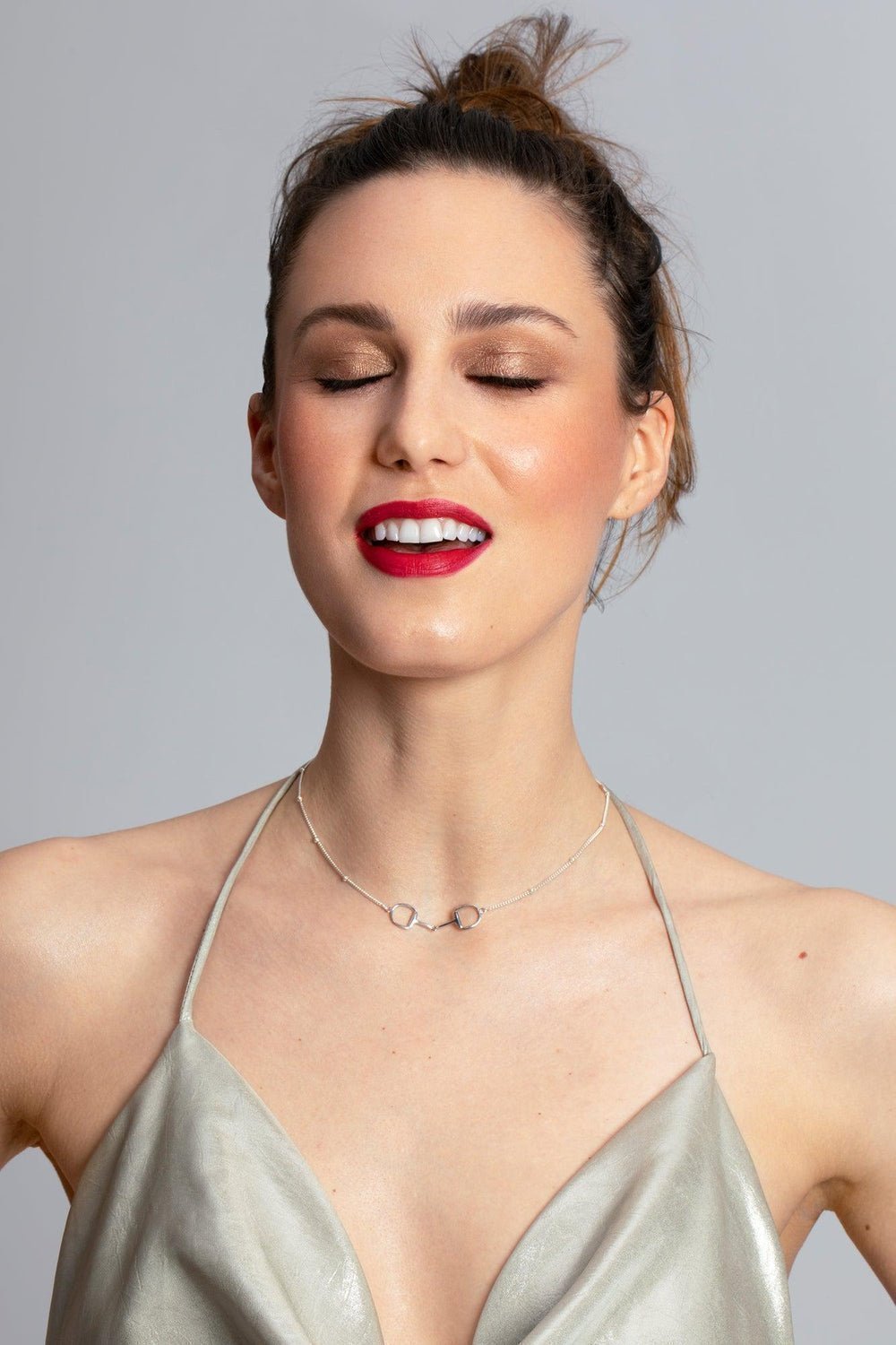 Brunet model woman with red lipstick and smiling, she wear a silver top.  She is wearing a stylish gold Snaffle Bit Necklace embodying sophistication in its design. The necklace chain  has a series of small, rounded beads interspersed along its length. This piece captures the essence of equestrian grace. Perfect for horse enthusiasts and fashion lovers, it adds a refined touch to any outfit and makes a memorable equestrian gifts for women and horse themed gifts.