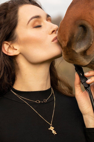 Brunet woman model kissing a horse, she is wearing a stylish gold horse necklace with a layering of Snaffle Bit necklace, the necklace chain is also gold and has a series of small rounded beads interspersed. This Snaffle Bit pendant necklace is the perfect for horse enthusiasts and fashion lovers, it adds a refined touch to any outfit and makes a memorable equestrian gifts for women and horse themed gifts, gold girls.