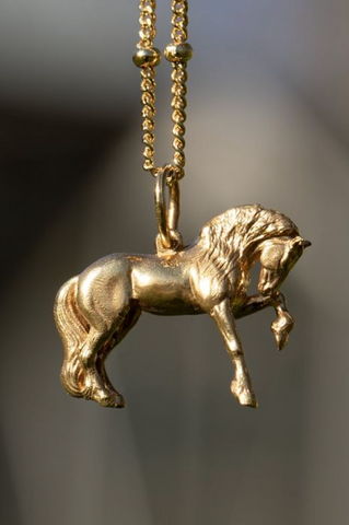 The Friesian Horse Pendant is a gold pendant necklace. The pendant is shaped like a horse in mid-stride, with detailed features such as its mane and tail. The necklace chain is also gold and has a series of small, rounded beads interspersed along its length. Capturing the powerful elegance and flowing grace of the Friesian horse  this is the perfect horse gift for friesian horses fans.