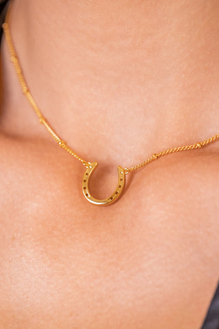 Close up of an elegant gold necklace adorned with a horseshoe charm, representing good fortune and sophistication. Our gold horseshoe bobble necklace is the perfect equestrian themed gift and as a good luck charm, horse lovers. The necklace chain is also gold and has a series of small, rounded beads interspersed along its length.  