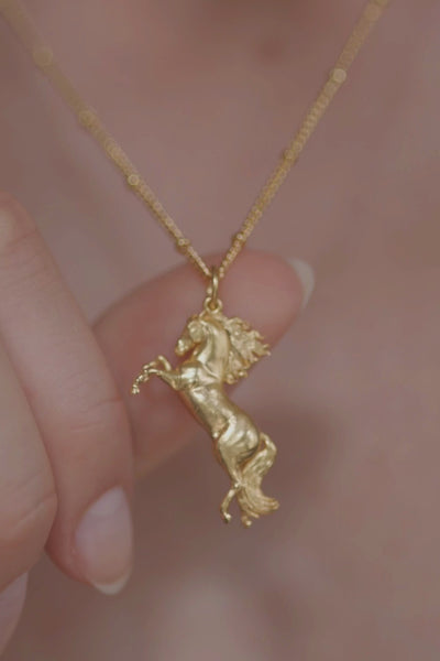 A beautifully crafted gold horse pendant, distinguished by its long tail and exquisite design. Our Rearing Wild Horse Pendant on a chain, highlighting the beauty of equestrian-inspired jewelry with  details on its  hair and tail. The necklace chain is also gold and has a series of small rounded beads interspersed. The Gold Wild Horse Pendant is the best gifts for an equestrian, horse riding, horse girls, cowgirls and for horse enthusiasts.
