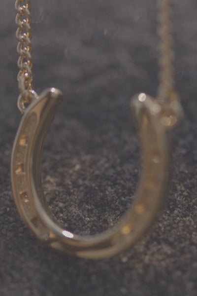 stylish gold necklace with a charming horseshoe pendant, embodying luck and refined beauty. The necklace chain is also gold and has a series of small, rounded beads interspersed along its length. The background appears to be a light-colored surface stone. This horse shoe necklace is the perfect equestrian themed gift and as a good luck charm, horse lovers.