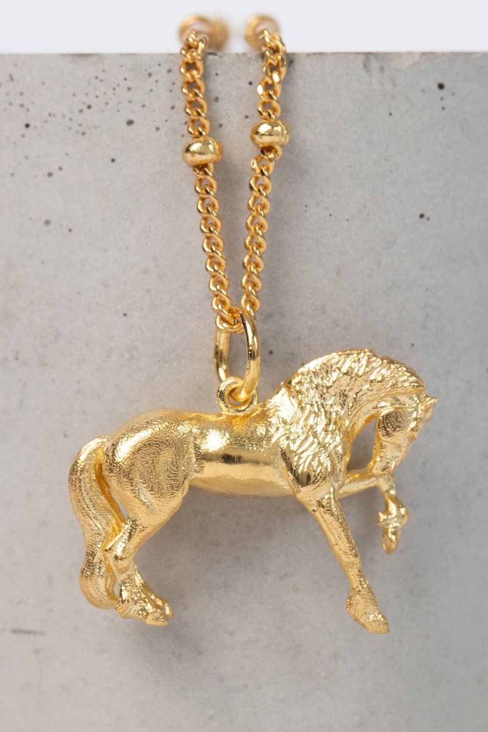 The Friesian Horse Pendant is a gold pendant necklace. The pendant is shaped like a horse in mid-stride, with detailed features such as its mane and tail. The necklace chain is also gold and has a series of small, rounded beads interspersed along its length. The background appears to be a plain or stone , light-colored surface. Capturing the powerful elegance and flowing grace of the Friesian horse  this is the perfect horse gift for friesian horses fans.