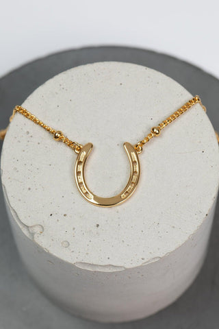 A stylish gold necklace with a charming horseshoe pendant, embodying luck and refined beauty. The necklace chain is also gold and has a series of small, rounded beads interspersed along its length. The background appears to be a light-colored surface stone. This horse shoe necklace is the perfect equestrian themed gift and as a good luck charm, horse lovers.