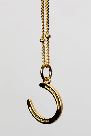 Discover our  equestrian inspired jewelry with the Gold  Horse Shoe Pendant Necklace. A gold necklace featuring a delicate horseshoe charm, symbolizing luck and elegance in a stylish design. 
The necklace chain is also gold and has a series of small, rounded beads interspersed along its length . The background appears to be a light-colored surface stone Crafted from high-quality gold is ideal for equestrians and horse life fans, gold jewelry girls.