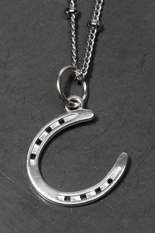 Discover our equestrian inspired jewelry with the Silver Horse Shoe Pendant Necklace. A silver necklace featuring a delicate horseshoe charm, symbolizing luck and elegance in a stylish design. 
The necklace chain is also silver and has a series of small, rounded beads interspersed. The background appears to be a light-colored surface stone Crafted from high-quality silver  is ideal for equestrians and horse life fans, silver jewelry girls.