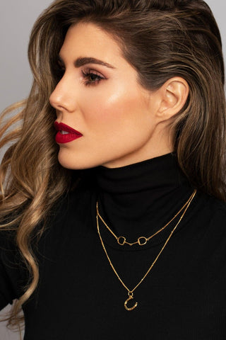 Brunet woman model with red lipstick and black sweater is wearing a stylish gold horseshoe necklace with a layering of Snaffle Bit necklace, emphasizing personal style and sophistication. A Gold horseshoe necklace with detailed real lifesize horseshoe features.  The necklace chain is also gold and has a series of small. This horseshoe pendant necklace is the perfect horse and equestrian themed gift and a good luck charm. Perfect for horse fans, pony lovers, show jumping, equestrian life.