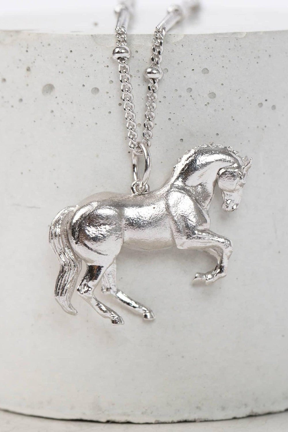 A silver horse pendant elegantly displayed, showcasing its intricate design and craftsmanship.The pendant is shaped like a wild horse in mid-stride, with detailed features such as its mane and tail.  The background appears to be a plain or stone , light-colored surface.  Perfect for horse lovers, fans of equestrian inspired jewelry, horseback riders, silver horse jewelry, unique equine gifts, horse girls. A dynamic piece of Horse Jewelry.