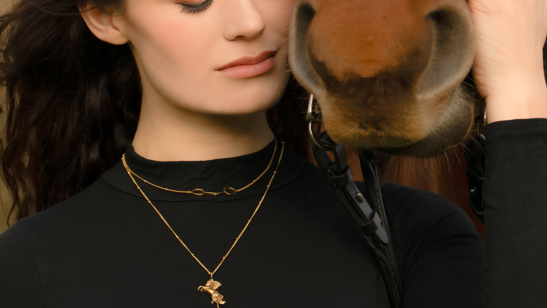 Brunet woman model with  horse, she is wearing a stylish Rearing Wild Horse Pendant with a layering of Snaffle Bit necklace, the necklace chain is also gold and has a series of small rounded beads interspersed. This  Rearing Wild Horse Pendant is the perfect for horse enthusiasts and fashion lovers, it adds a refined touch to any outfit and makes a memorable equestrian gifts for women and horse themed gifts, gold girls.
