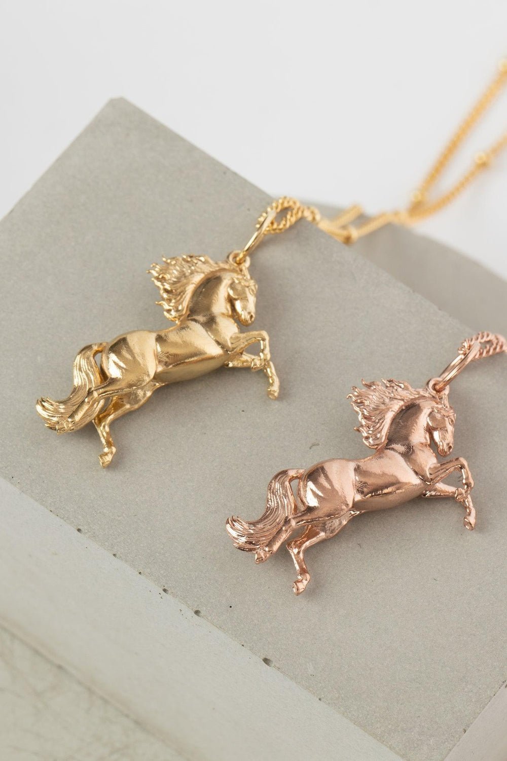 Two gold rearing wild horse pendant on a chain, highlighting the beauty of equestrian-inspired jewelry with  details on its  hair and tail. The  Gold Wild Horse Pendant is the best gifts for an equestrian, horse riding, horse girls. This masterpiece offers both beauty and freedom symbolism in one. Stone or marble background.