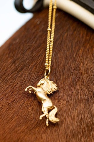 A beautifully crafted gold-plated horse pendant, distinguished by its long tail and exquisite design. Our Rearing Wild Horse Pendant on a chain, highlighting the beauty of equestrian-inspired jewelry with  details on its  hair and tail. The necklace chain is also gold and has a series of small rounded beads interspersed. The Gold Wild Horse Pendant is the best gifts for an equestrian, horse riding, horse girls, cowgirls and for horse enthusiasts.
A brown horse's neck in the background.