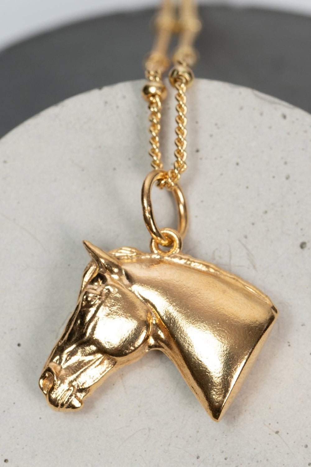 A gold horse head necklace with detailed features such as its mane. The necklace chain is also gold and has a series of small, rounded beads interspersed along its length. 
The gold horse head pendant is elegantly displayed on a pristine white marble surface. Ideal as a unique gift, this pendant celebrates the beauty and strength of horses. Best gifts for horseback riders and one of a kind horse gift ideas for her.