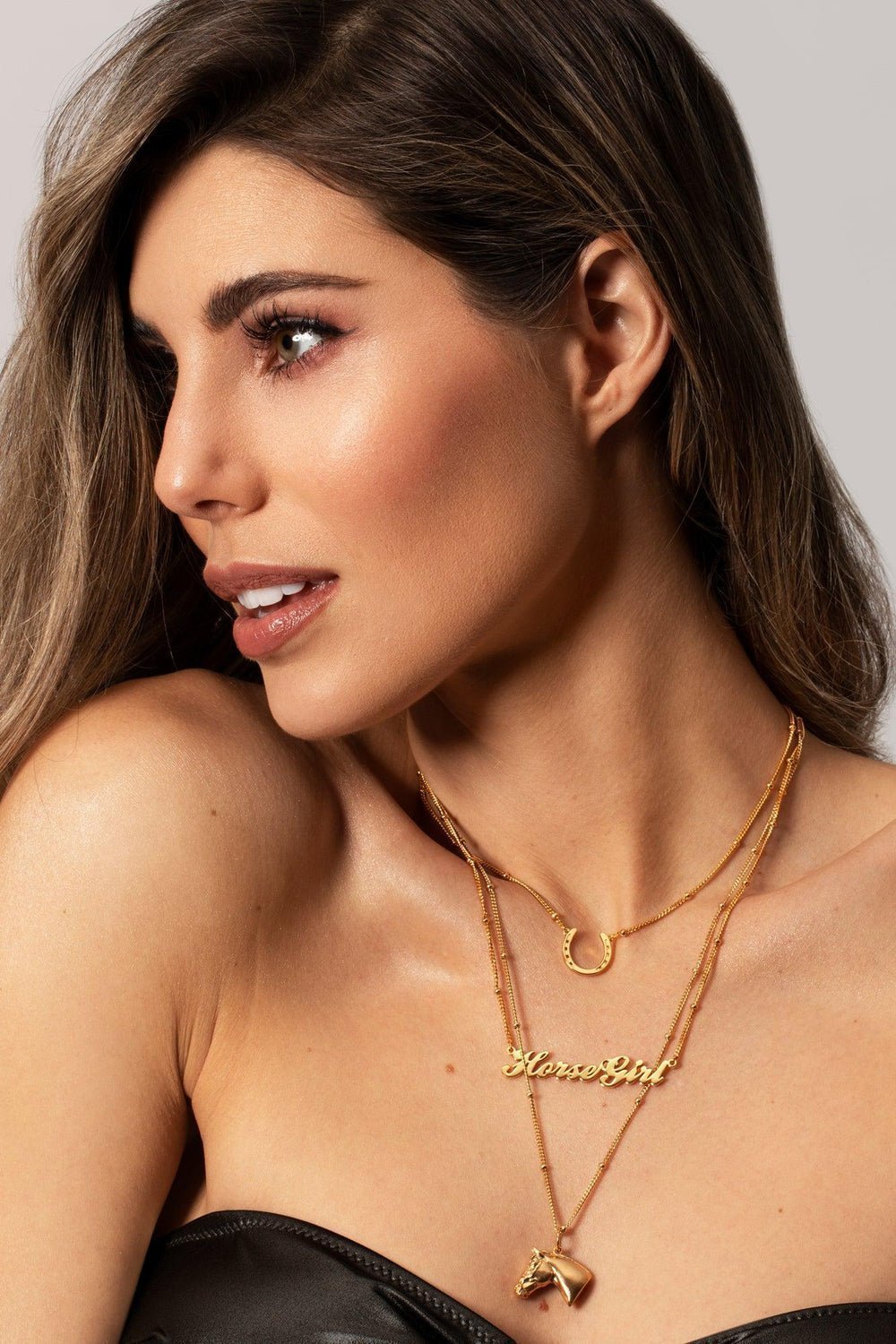 A brunet woman model displays a stylish gold horse head necklace layering with a name pendant necklace: horse girl and also horseshoe necklace, emphasizing personal style and sophistication. A Gold horse head necklace with detailed features such as its mane. The necklace chain is also gold and has a series of small. Ideal as a unique gift, this pendant celebrates the beauty and strength of horses. Best gifts for horseback riders and one of a kind horse gift ideas for her. A unique horse gift and equestrian 