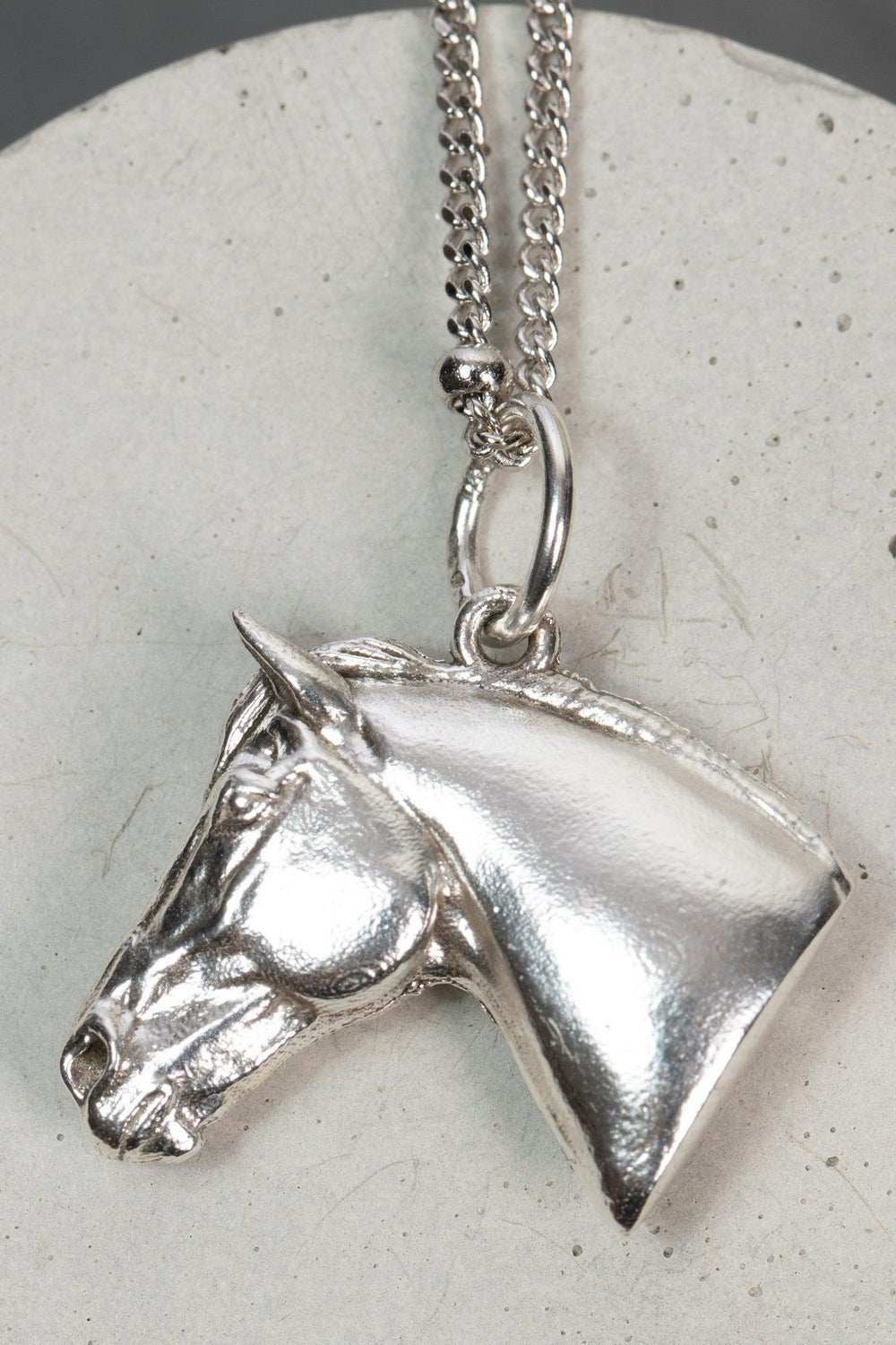 A silver horse head necklace with detailed features such as its mane. The necklace chain is also silver  and has a series of small, rounded beads interspersed along its length. The silver horse head pendant is elegantly displayed on a pristine white marble surface. Ideal as a unique gift, this pendant celebrates the beauty and strength of horses. Best gifts for horseback riders and one of a kind horse gift ideas for her.